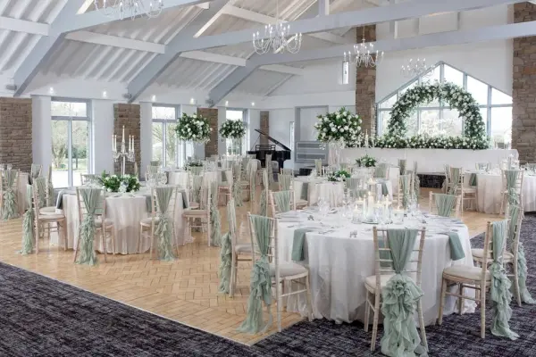 Elegant reception space featuring round tables adorned with pale green sashes, lush floral arrangements, and large windows for natural light.