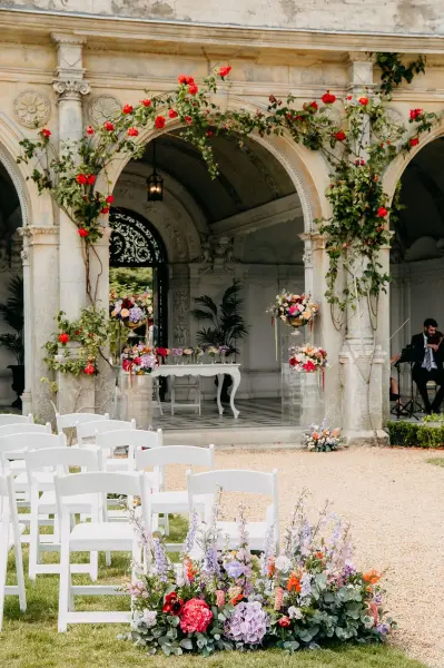 Charming outdoor ceremony space featuring elegant arches adorned with greenery and vibrant flowers, set with white chairs and a decorative table.