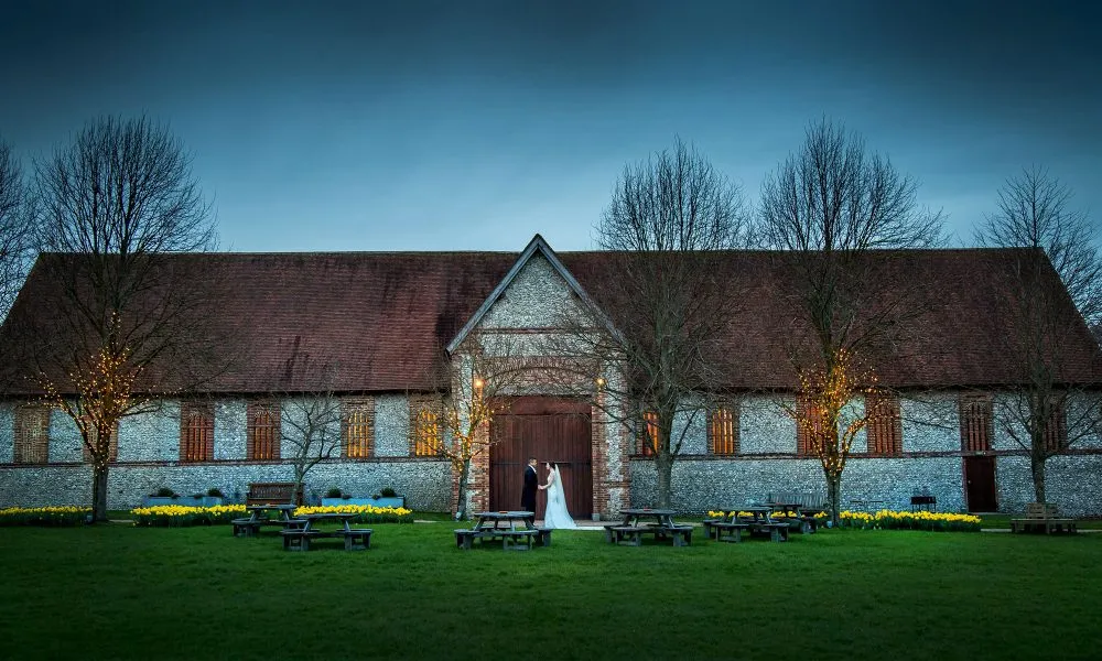 Charming rustic barn venue, with a stone facade and warm lights, surrounded by grassy grounds and yellow flowers, perfect for outdoor ceremonies.