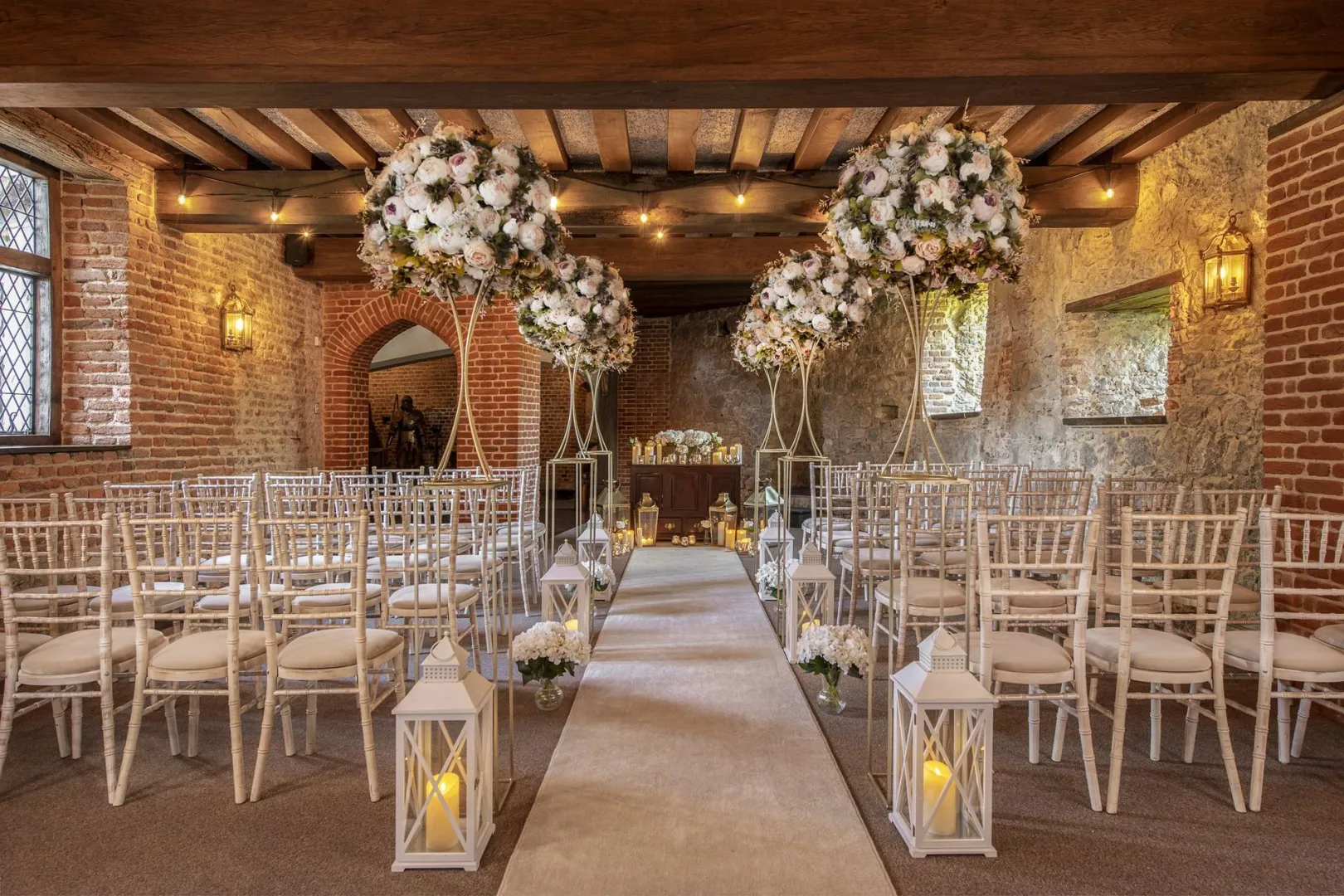 Charming indoor ceremony space featuring white Chiavari chairs, floral arrangements, and soft lighting, set against rustic stone walls.