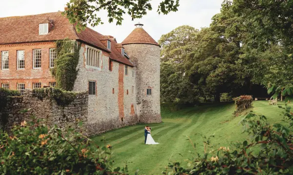 A couple embraces on a lush green lawn, framed by a historic stone building with a charming turret and surrounding trees.