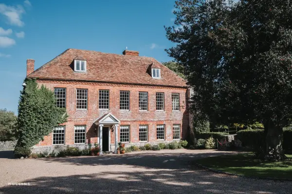 Charming brick estate with large windows surrounded by lush greenery and a gravel driveway, perfect for outdoor ceremonies or receptions.