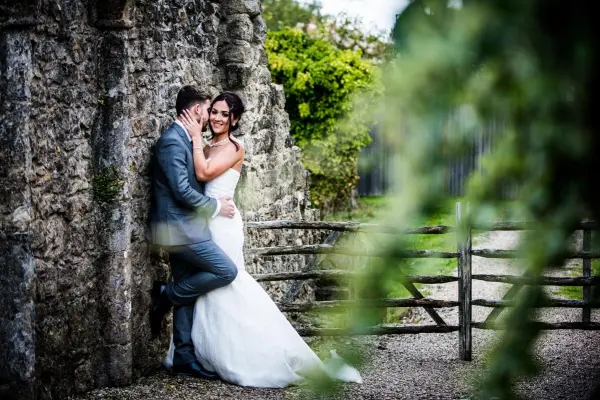 Couple embracing against a rustic stone wall, surrounded by greenery and a charming wooden gate, creating a romantic wedding moment.