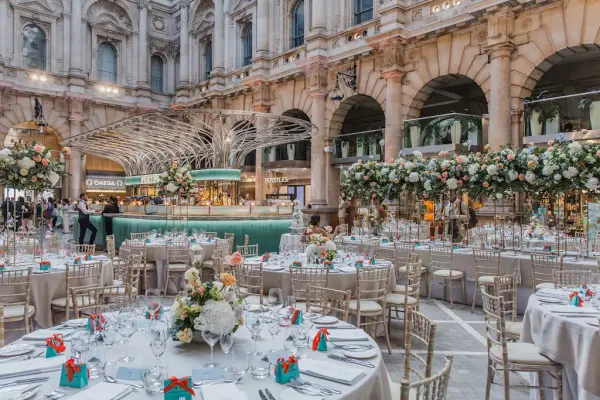 Elegant dining setup with round tables adorned with floral centerpieces, gold chairs, and lush greenery in a grand, sophisticated venue.