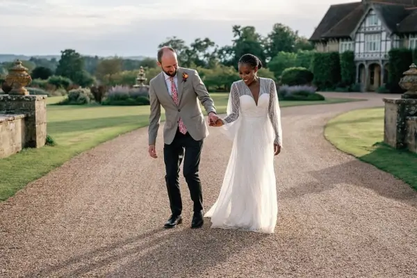 A couple strolls hand-in-hand along a scenic gravel path, surrounded by lush gardens and an elegant venue in the background.