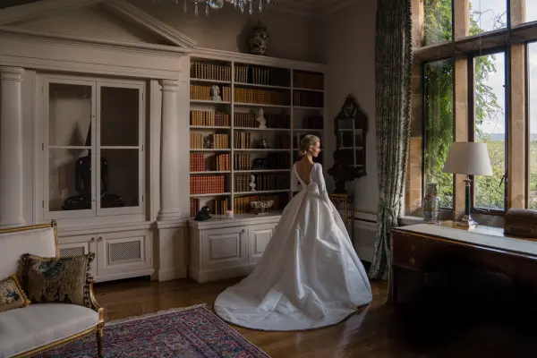 A bride in a stunning gown stands gracefully in an elegantly decorated room with large windows, wooden floors, and a cozy library backdrop.