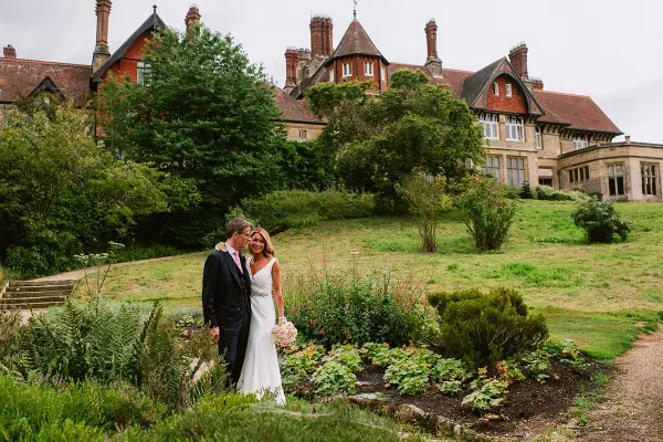 A couple embraces in a lush garden, framed by a grand historic mansion with elegant architecture and manicured greenery.