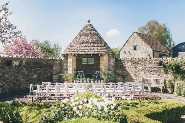 Charming outdoor ceremony space featuring rustic decor, a stone gazebo, and wooden seating surrounded by vibrant greenery and blooms.