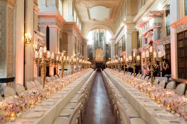 Elegant dining setup in a grand hall, featuring long tables adorned with pink floral centerpieces and glowing candlelight under ornate architecture.