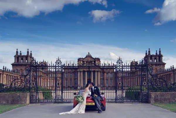 A couple shares a kiss in front of a grand estate, featuring ornate gates and majestic architecture against a bright blue sky.