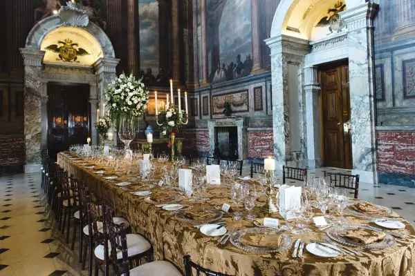 Elegant dining setup in a grand hall, featuring a long table adorned with gold linens, fine china, and lush floral arrangements.