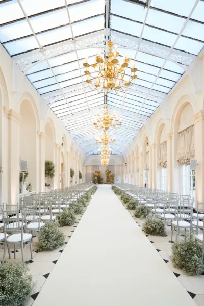 Bright glass-enclosed ceremony space featuring elegant chandeliers, lush greenery, and rows of silver chairs lining a soft white aisle.