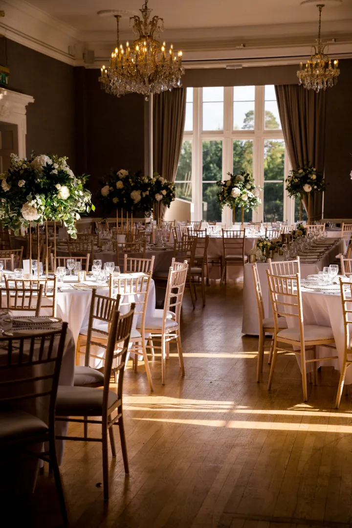 Elegant reception space featuring golden chandeliers, tall floral arrangements, and sunlit wooden floors, perfect for a romantic wedding celebration.