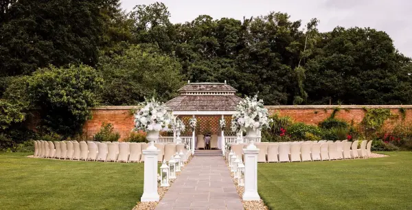 A charming outdoor ceremony space featuring a floral-adorned gazebo, lined with white chairs on a manicured lawn surrounded by greenery.