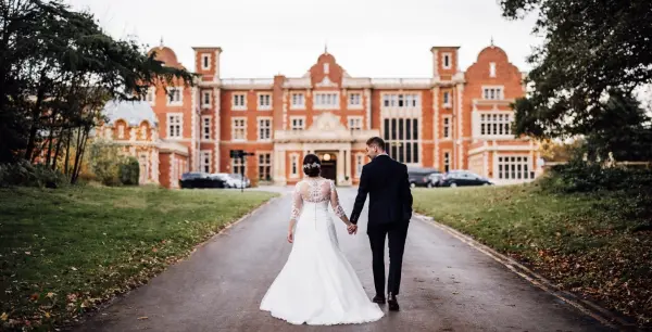 A bride and groom walk hand-in-hand down a tree-lined path, framed by an elegant red-brick mansion in a lush, romantic setting.