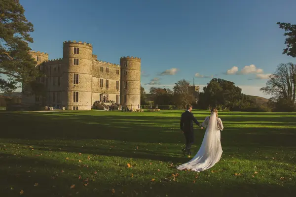Couple walking hand in hand across a lush green lawn with a stunning castle backdrop, perfect for a romantic outdoor wedding ceremony.