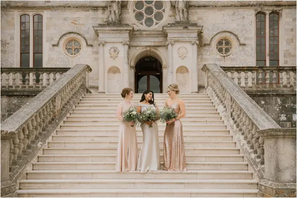 Three bridesmaids in elegant attire holding lush bouquets stand on grand stone steps leading to a majestic, vintage venue entrance.
