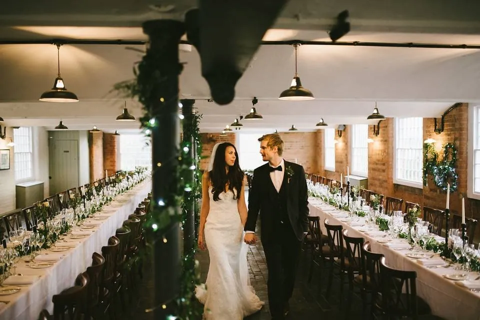 A couple walks hand in hand through an elegant reception space featuring long tables adorned with greenery and soft overhead lighting.