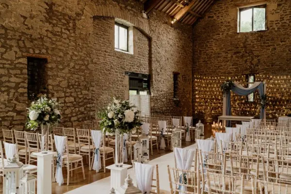 Charming rustic barn venue featuring stone walls, elegant chairs, floral arrangements, and a stylish ceremony arch for intimate weddings.