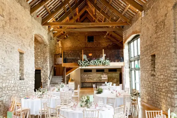 Rustic barn venue featuring high wooden beams, stone walls, and elegantly set round tables adorned with floral centerpieces.