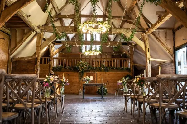 Charming rustic barn interior with wooden beams, elegant chandeliers, and flower-adorned chairs set for a wedding ceremony.