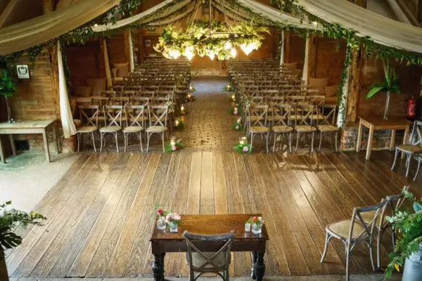 Rustic indoor wedding venue featuring wooden floors, crossback chairs, a floral canopy, and warm, inviting lighting for a charming ceremony.