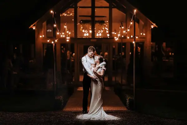 Couple embracing in front of a beautifully lit venue, featuring warm string lights and inviting glass doors, perfect for evening celebrations.