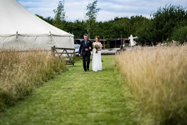 A couple walks down a grassy path lined with tall grass, leading to a charming marquee amidst a rustic outdoor setting.