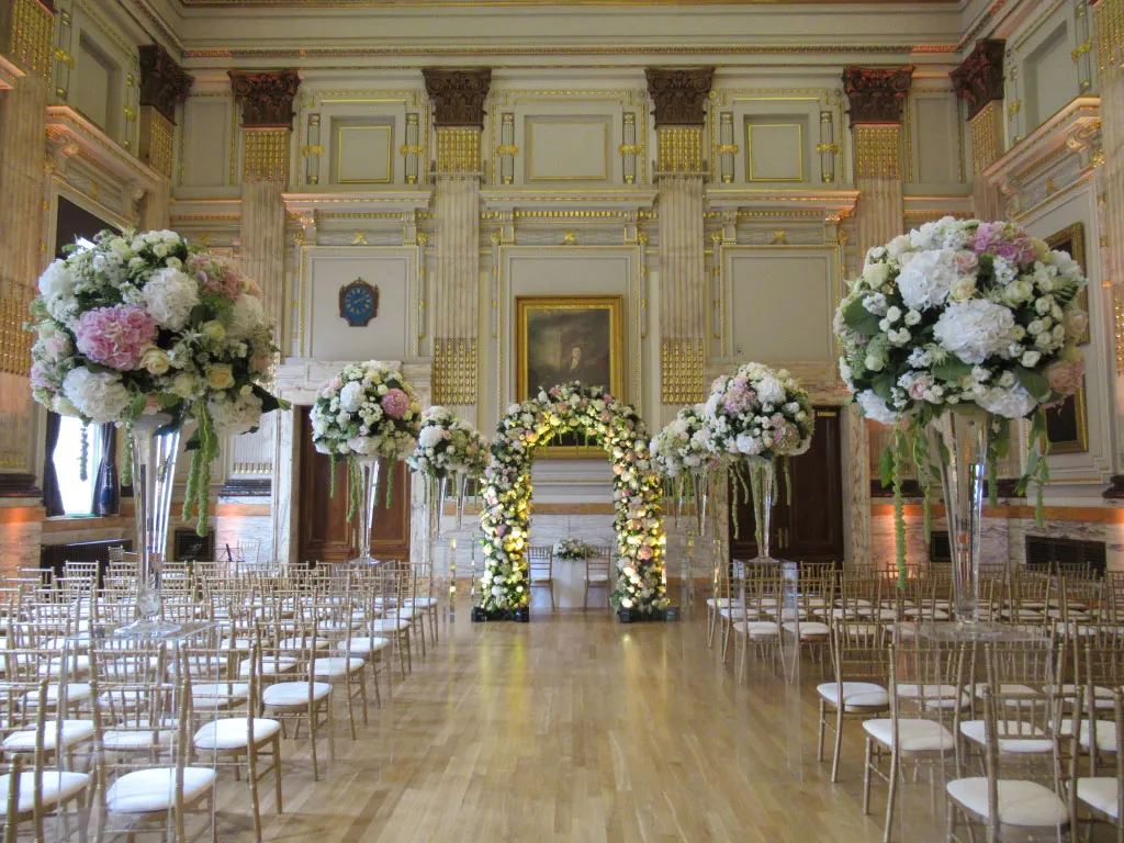 Elegant ballroom featuring floral arrangements, a stunning floral arch, and rows of golden chairs for a beautiful wedding ceremony.