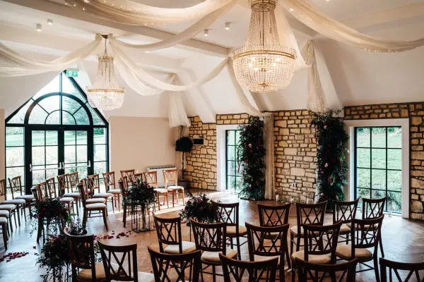 Charming indoor ceremony space featuring wooden chairs, lush greenery, elegant chandeliers, and soft draped fabric over a stone wall.