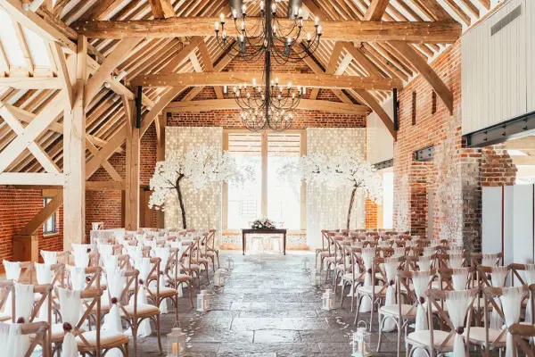 Rustic barn wedding venue featuring elegant wooden beams, a chandelier, and decorated with white florals, offering a charming ceremony space.
