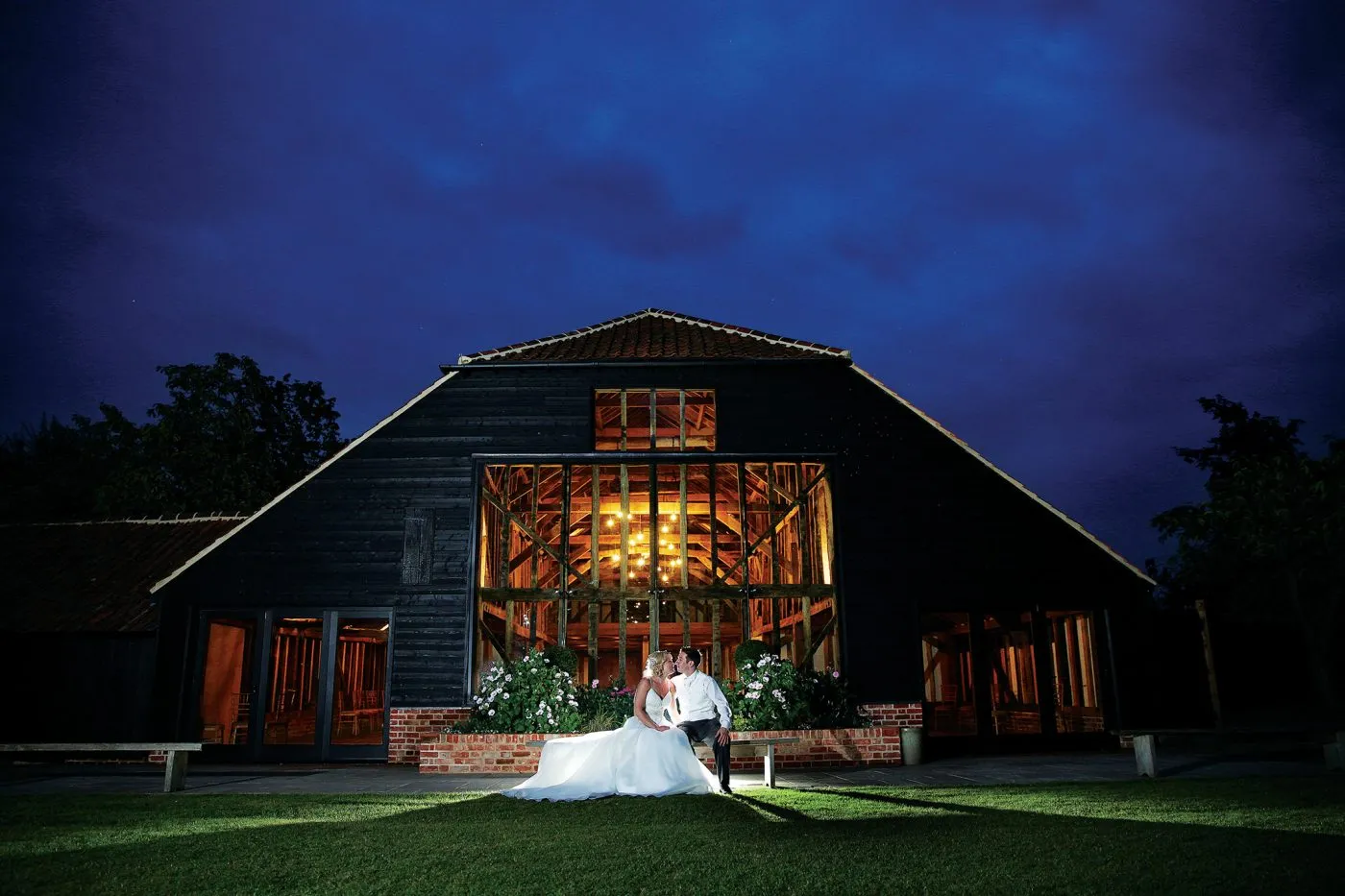 A couple sits together in front of a rustic barn venue, featuring large windows illuminated against a twilight sky. Lush greenery frames the scene.