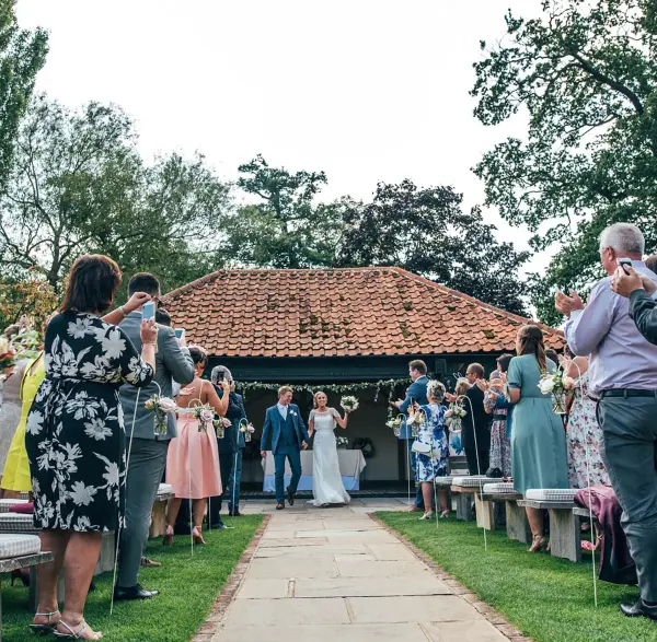 Charming outdoor ceremony with guests applauding, elegant stone path, and a scenic backdrop of lush greenery and a quaint structure.