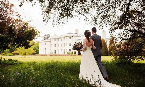 Elegant couple walking hand in hand through lush greenery, with a grand estate in the background, ideal for romantic outdoor ceremonies.