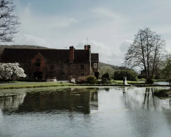 Charming historic venue by a serene pond, featuring lush gardens and a rustic red brick building, perfect for an outdoor wedding.