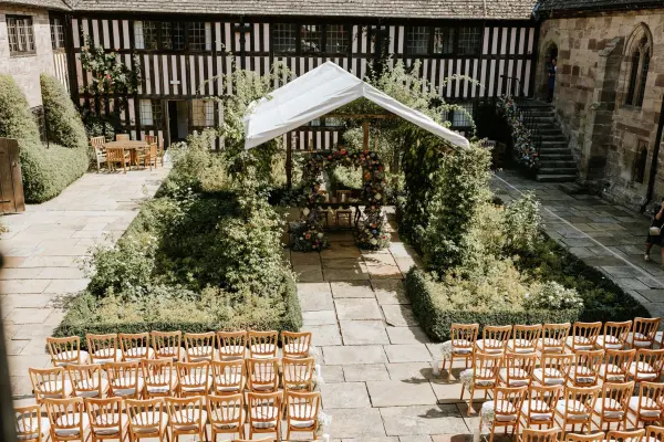 Charming outdoor ceremony space with wooden chairs, lush greenery, and a floral arch beneath a white canopy, set in a historic courtyard.
