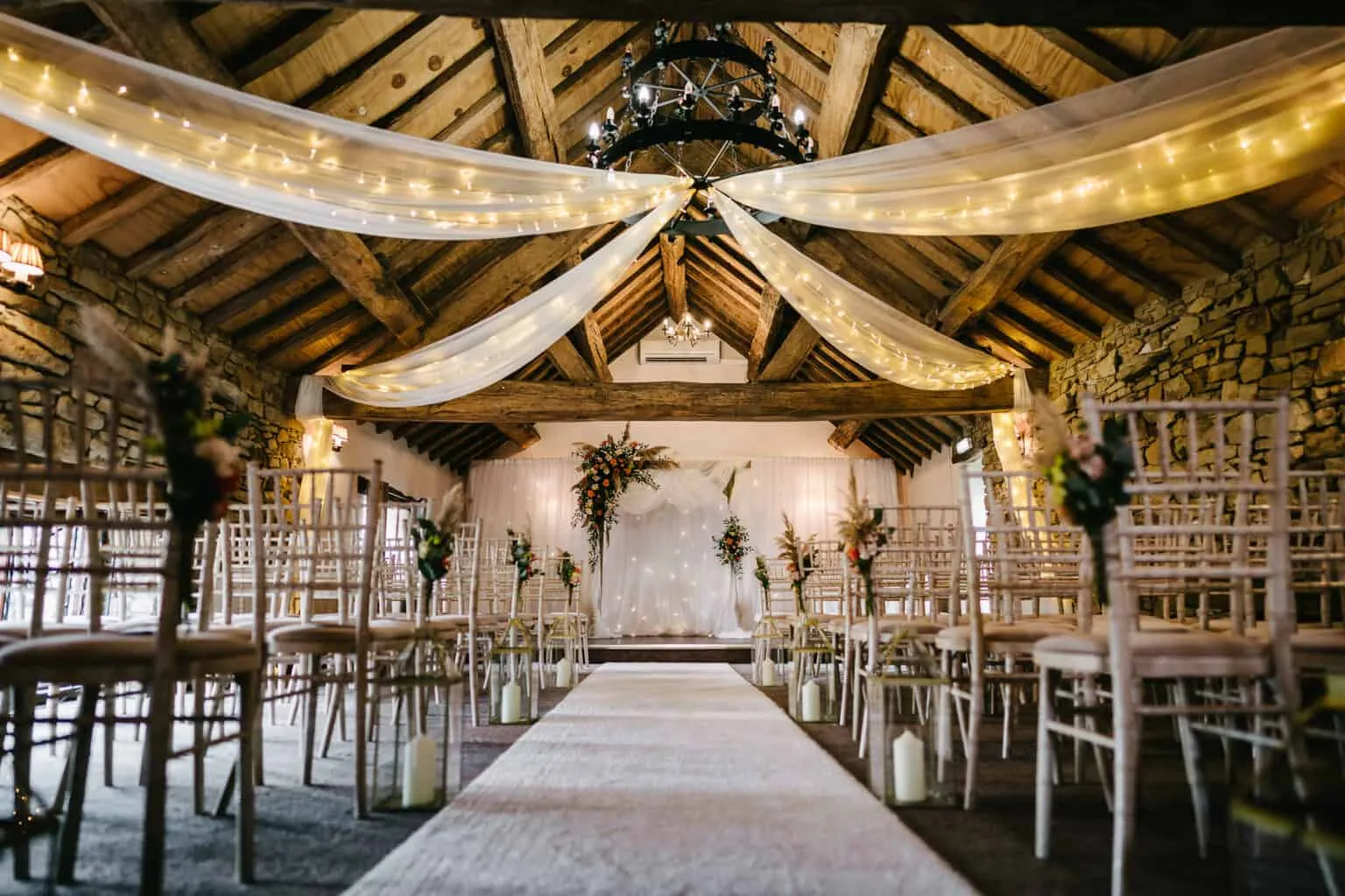 Charming rustic barn with wooden beams, twinkling fairy lights, and elegant ceremony setup featuring chiavari chairs and floral accents.