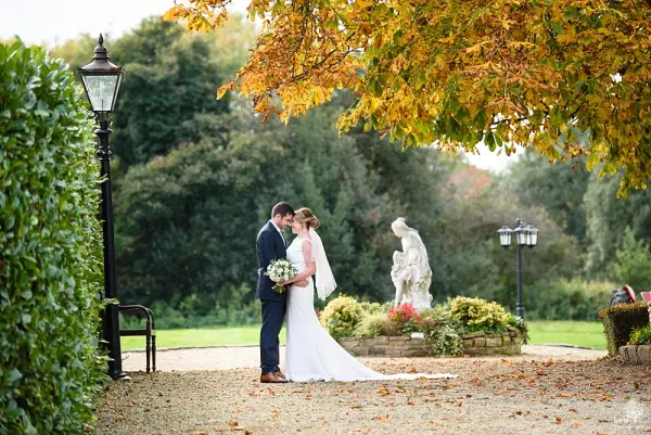 A couple shares a romantic kiss in a picturesque garden, framed by autumn leaves, charming lanterns, and a serene statue.