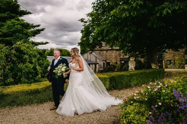 A bride in a flowing gown walks with her father, surrounded by lush greenery and charming stone buildings in a picturesque garden setting.