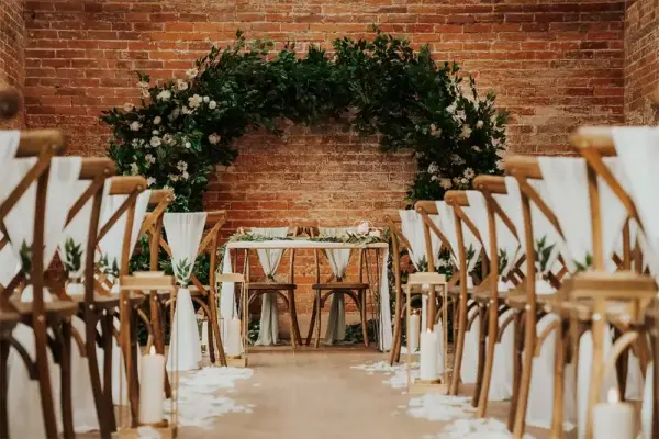 Charming indoor ceremony space featuring rustic wooden chairs, floral arch, and elegant decor against a brick backdrop.