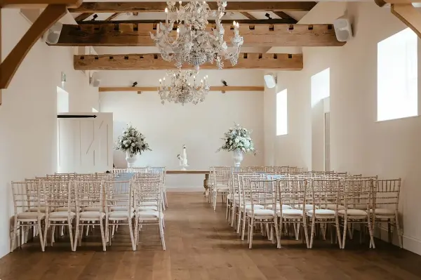 Elegant indoor ceremony space featuring a wooden beam ceiling, sparkling chandelier, and rows of chiavari chairs set for a wedding.