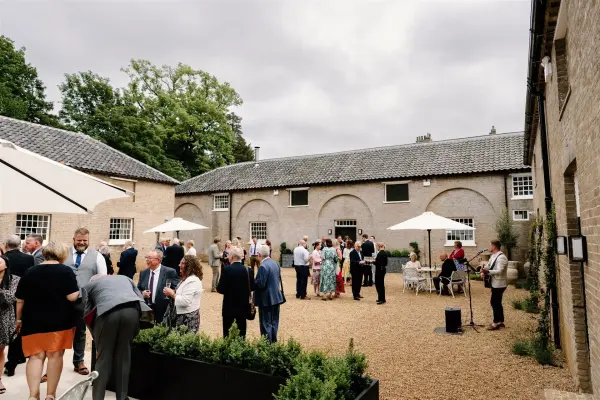 Charming outdoor courtyard with guests mingling under umbrellas, surrounded by rustic brick buildings and lush greenery. Perfect for a wedding.