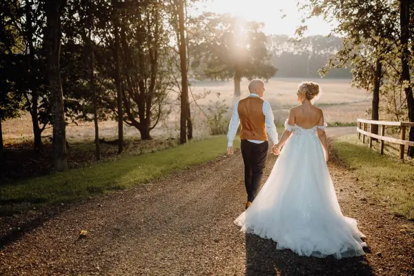 A couple walks hand-in-hand down a scenic path, flanked by trees, as golden sunlight filters through, creating a romantic outdoor setting.