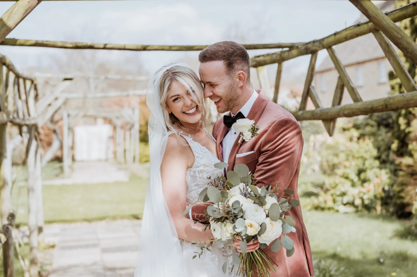 A joyful couple stands under a rustic wooden archway, surrounded by lush greenery, ready to embrace their outdoor wedding.
