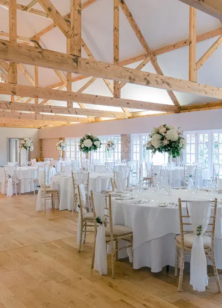 Spacious reception area featuring elegant round tables adorned with floral centerpieces and a rustic wooden beam ceiling.