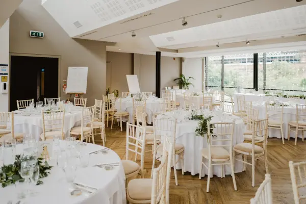 Bright, stylish event space featuring round tables with elegant white linens, chiavari chairs, and large windows overlooking greenery.