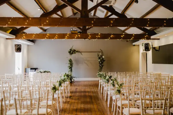 Elegant ceremony space featuring exposed wooden beams, fairy lights, and rows of chiavari chairs, with a floral arch as the focal point.