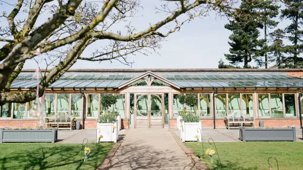 Charming glass-encased venue surrounded by lush greenery, leading to a serene outdoor ceremony space and inviting pathways.