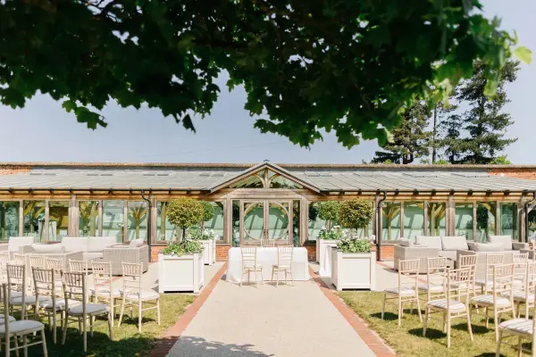 A charming outdoor ceremony space features elegant chairs, lush greenery, and a beautiful glass-enclosed structure for an intimate wedding atmosphere.