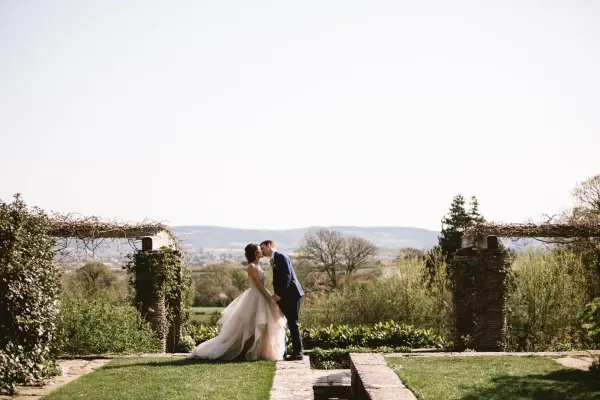 A couple shares a romantic kiss in a lush outdoor setting, framed by stone pillars and green foliage, overlooking serene hills.